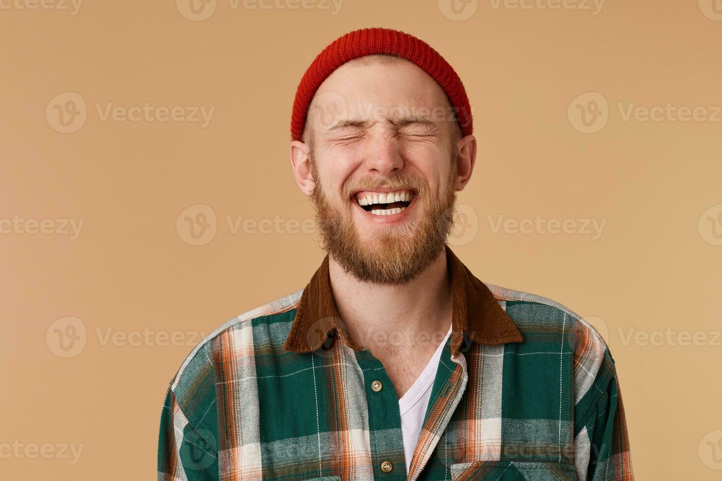 Laughing man in checkered shirt and red hat over beige wall. Toothy smile and beard. Male laughing out loud at funny meme he found on internet. Positive human facial expressions and emotions photo