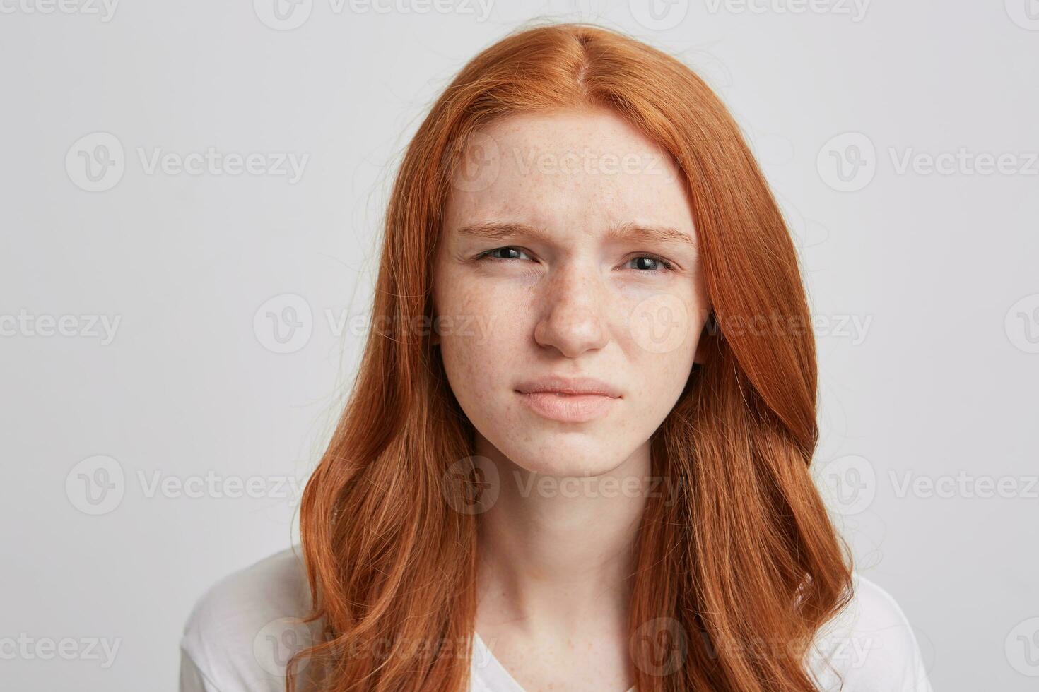 Portrait of sad unhappy young woman with long wavy red hair and freckles feels upset and looks directly in camera isolated over white background photo