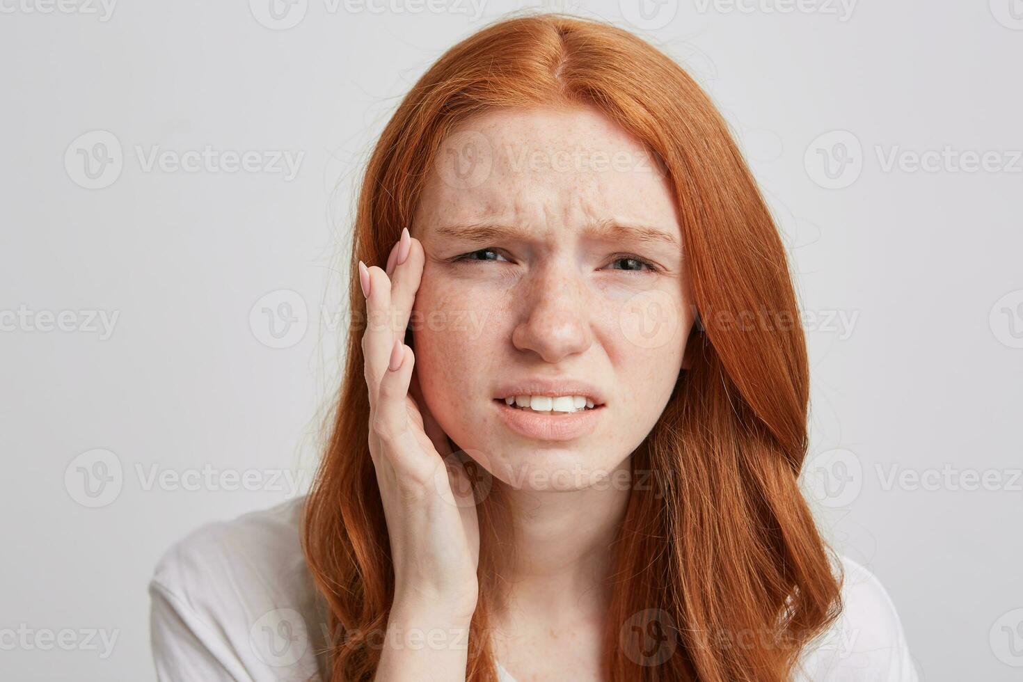 Closeup of upsed depressed young woman with long wavy red hair and freckles touching her temple and suffering from headache isolated over white background photo