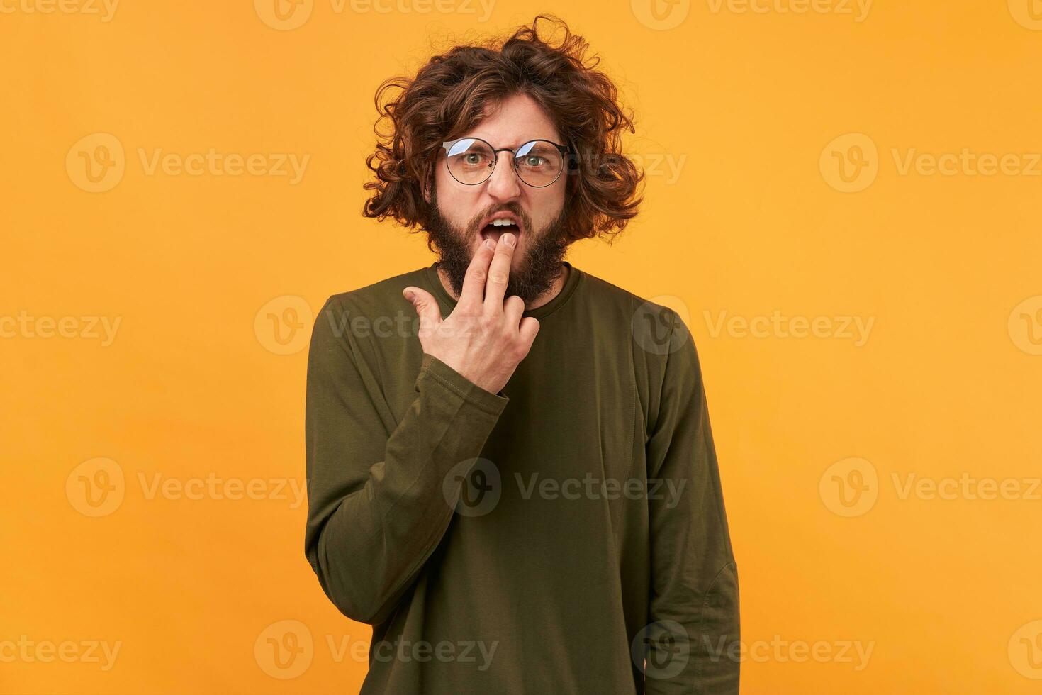 Attractive caucasian bearded male in glasses puts fingers in mouth showing nausea feels disgust about new work with unpleasant people. Facial expression. Isolated over yellow background photo