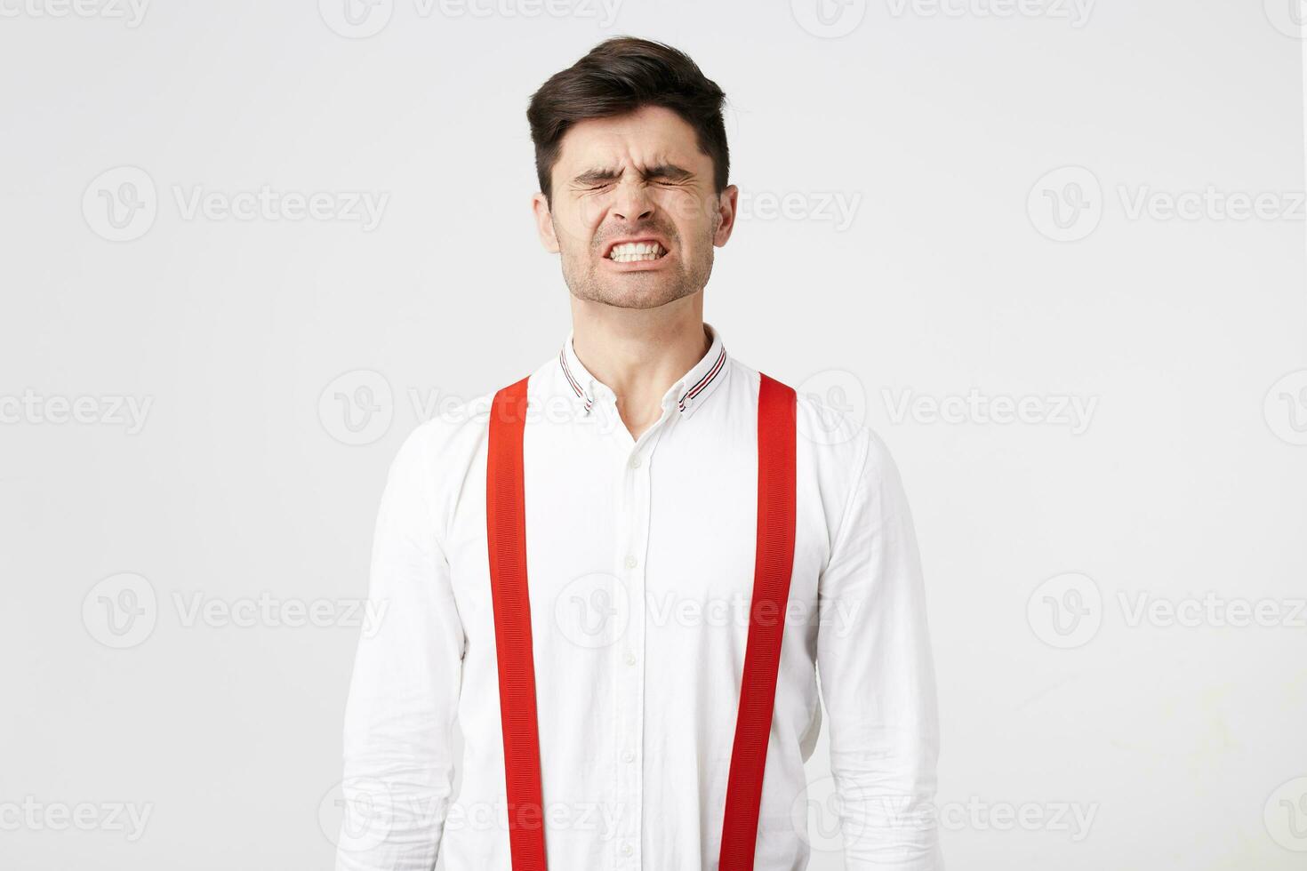 A man dressed in a white shirt and red suspenders, screw up his eyes,showing,squeezing white teeth as if he had lifted something heavy, an unbearable burden, severe pain,isolated over white background photo