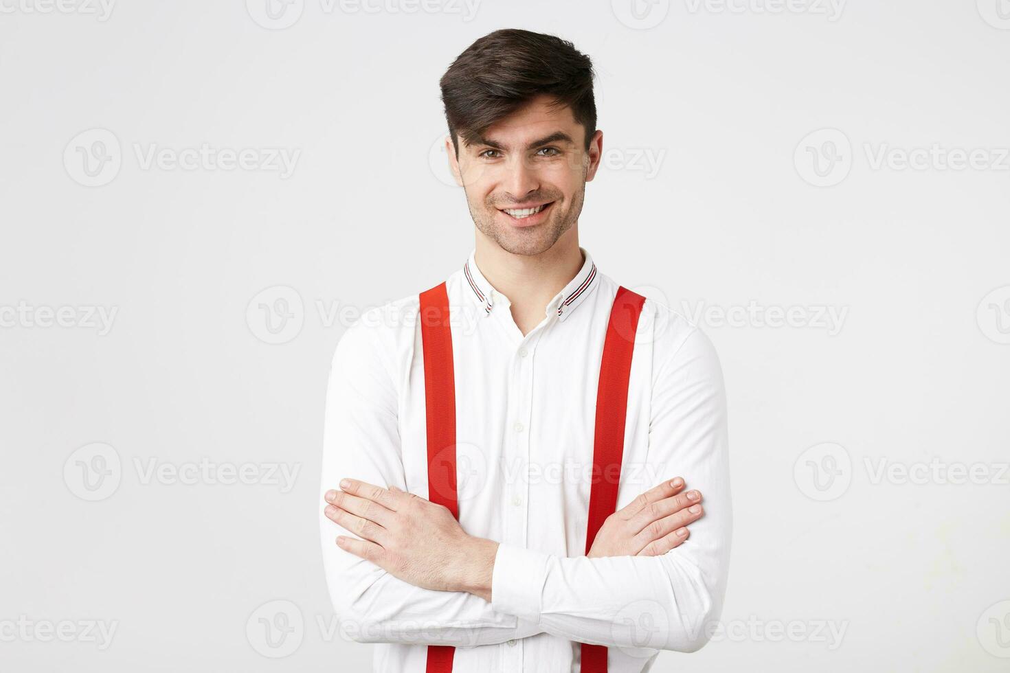 confidente joven hipster con oscuro pelo sin afeitar en pie con su brazos cruzado, vistiendo un blanco camisa, rojo tirantes, sonriente mira seguro de sí mismo y contento aislado en un blanco antecedentes. foto