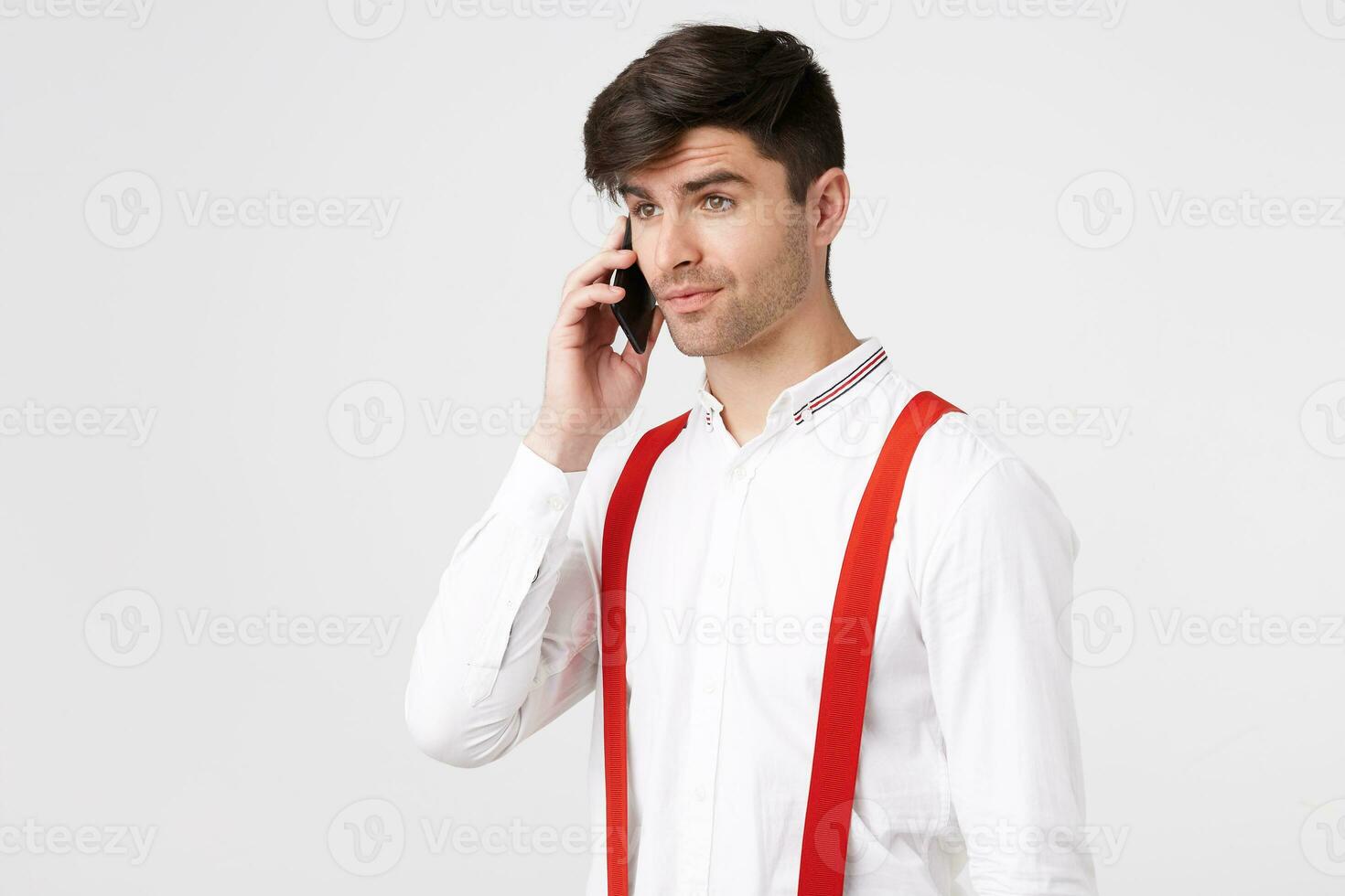 Attractive cute dark haired unshaven guy talking on a mobile phone looking the side feels a little surprised, phone conversation gives pleasure, wears shirt and red suspenders, on white background photo