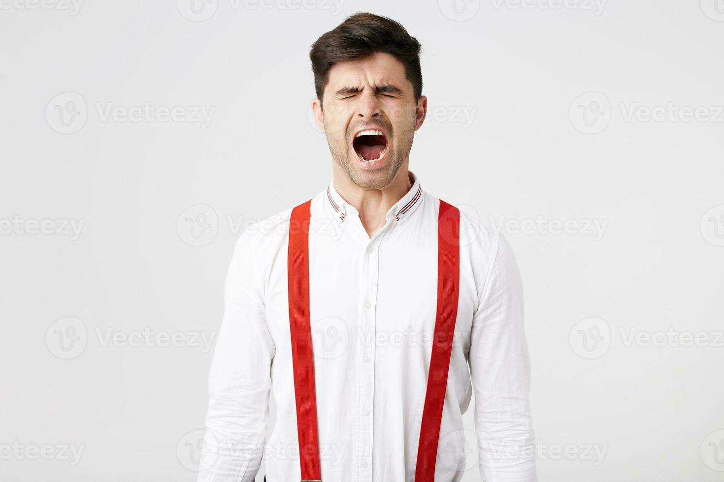 retrato de hermoso chico despierta arriba bostezos su ojos cerrado, vistiendo camisa y rojo tirantes tener no dormido cansado, difícil día, un lote de trabajar, querer café, boca es amplio abierto, aislado en un blanco antecedentes. foto