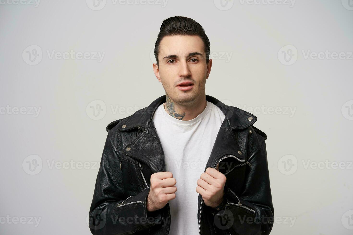 retrato de atractivo rebelde, brutal con un elegante Corte de pelo y peinado con un grasa, tatuaje en el cuello, mantiene manos me gusta poniendo en negro estilo rockero cuero chaqueta, terminado blanco antecedentes foto