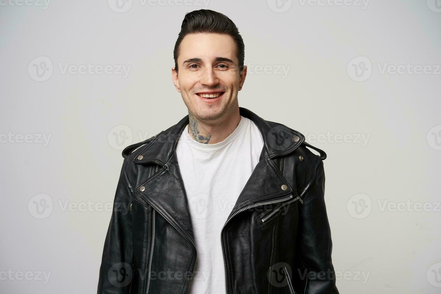 Attractively smiling young rebel, with a stylish hairstyle in briolin, tattoo on the neck, dressed in the style of 50s-60s in black leather jacket and white T-shirt, over white background photo