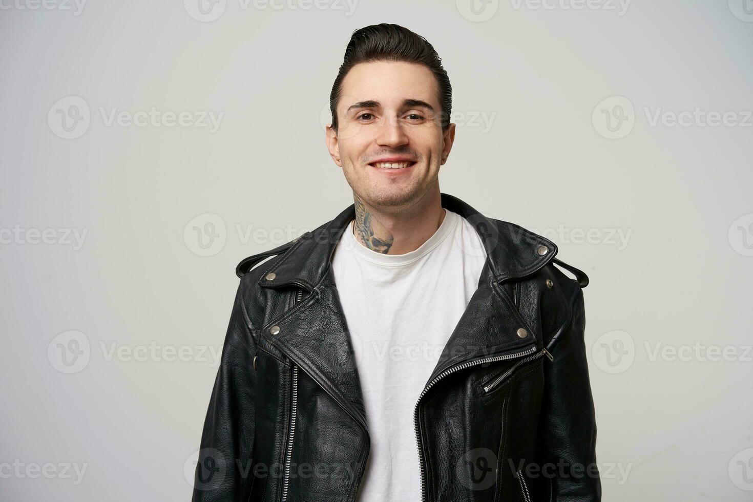 Charmingly, enticing, boldly smiling young rocker, rebel, motto racer with a stylish hairstyle in briolin, tattoo on the neck, dressed in the style of 50s-60s in black leather jacket, white background photo