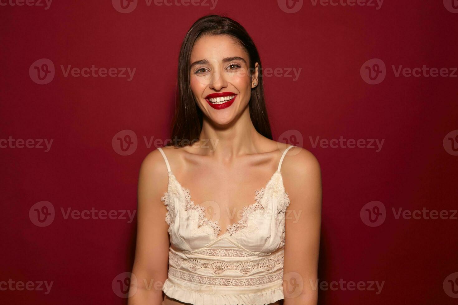 Happy young brown-eyed dark haired lady with evening makeup looking positively at camera with wide pleasant smile, standing over burgundy background in elegant wear photo