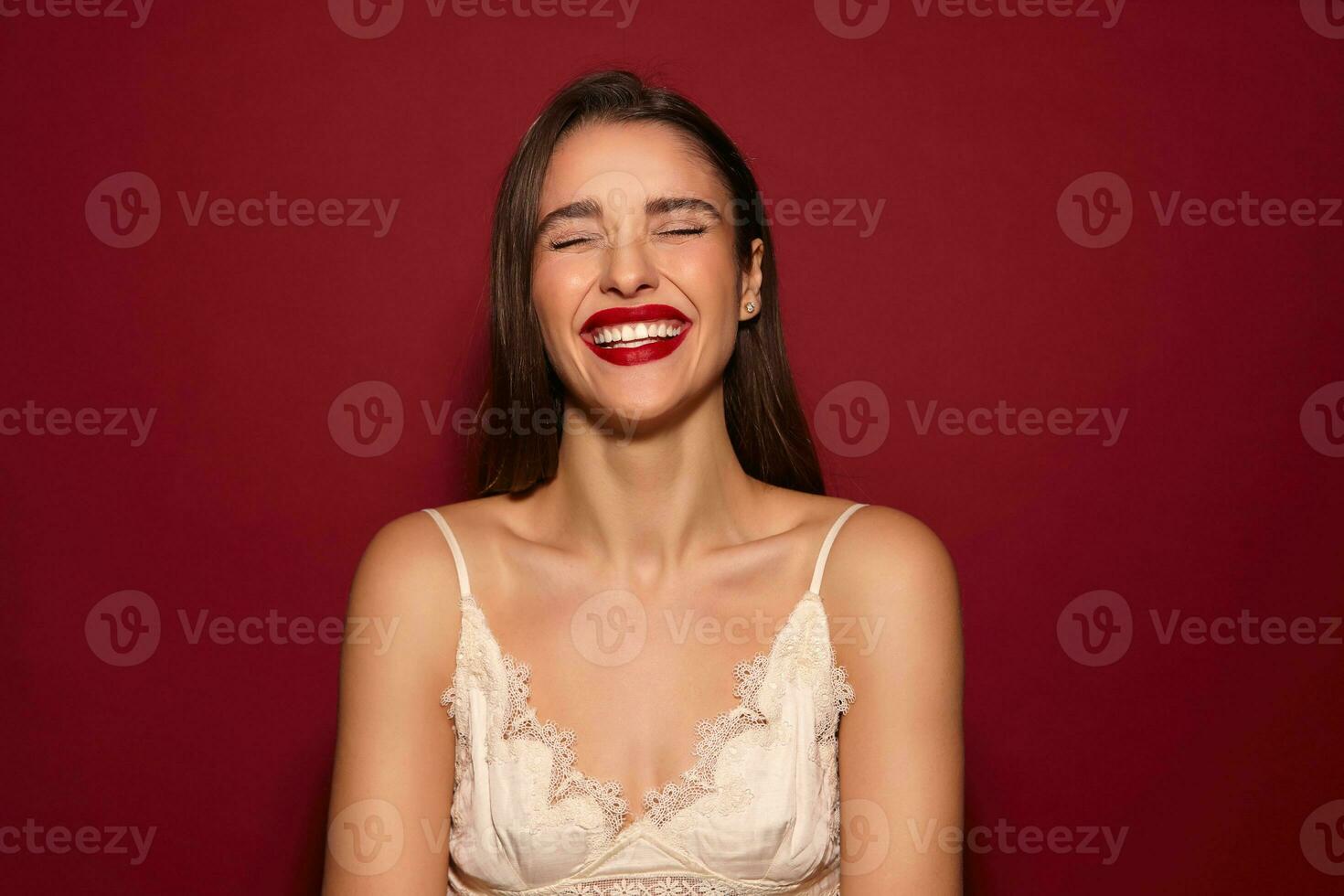 Pleasant looking young joyful dark haired lady with festive mua keeping her eyes closed while smiling happily, standing over burgundy background with hands down photo