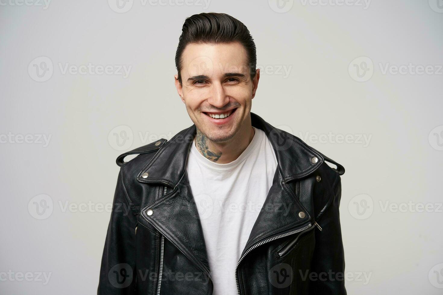 photo of attractively smiling young rocker man, rebel, motto racer, stylish hairstyle in briolin, looks in the style of 50s-60s, tattoo on the neck, in black leather jacket,over white background