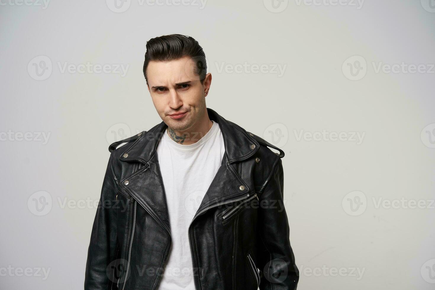 Young rocker, rebel, motto racer with a stylish hairstyle in briolin, tattoo on the neck, looking in the style of 50s-60s in black leather jacket, looks dissatisfied, purses one corner of the lips photo