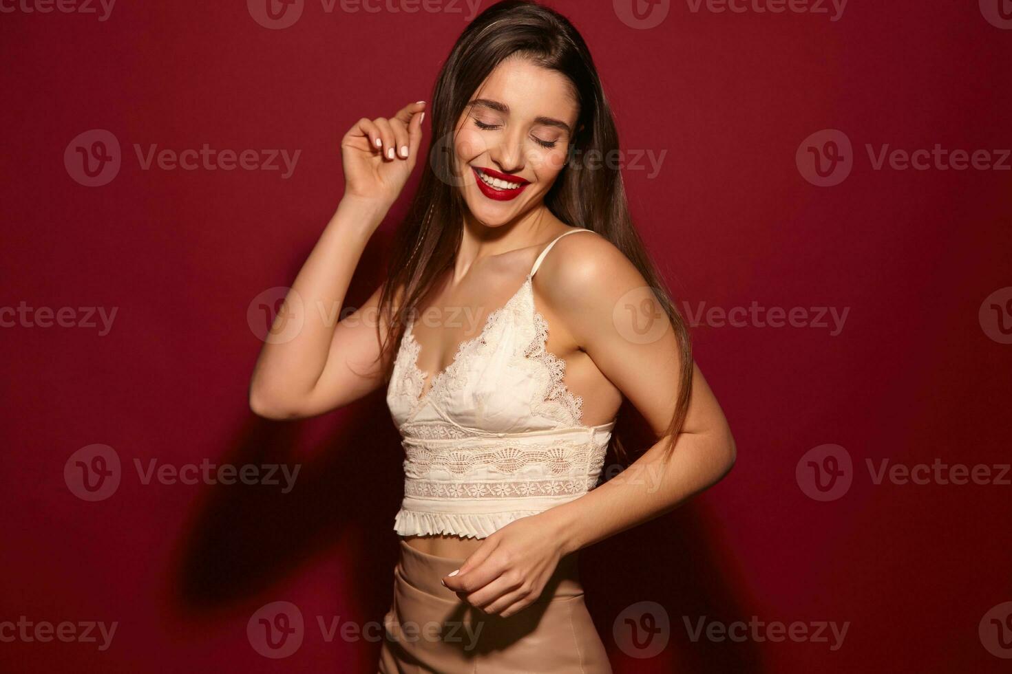 Good looking young lovely long haired brunette lady with red lips keeping her eyes closed and smiling cheerully, standing over burgundy background with raised hands photo