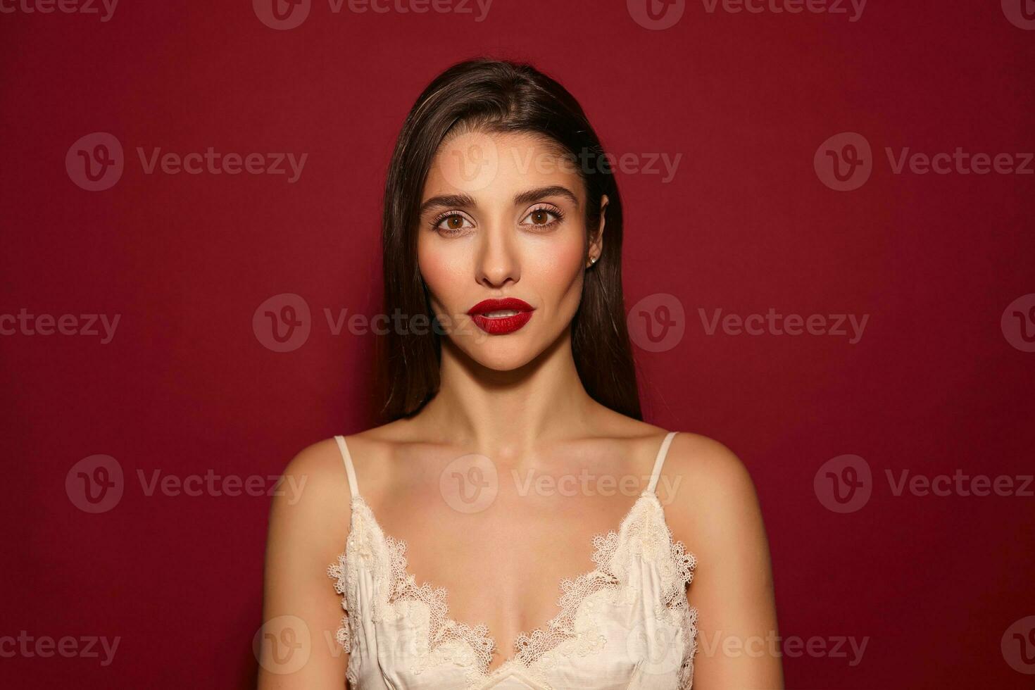 Portrait of young attractive brown-eyed brunette woman with evening makeup looking seriously at camera while posing over burgundy background in elegant wear photo