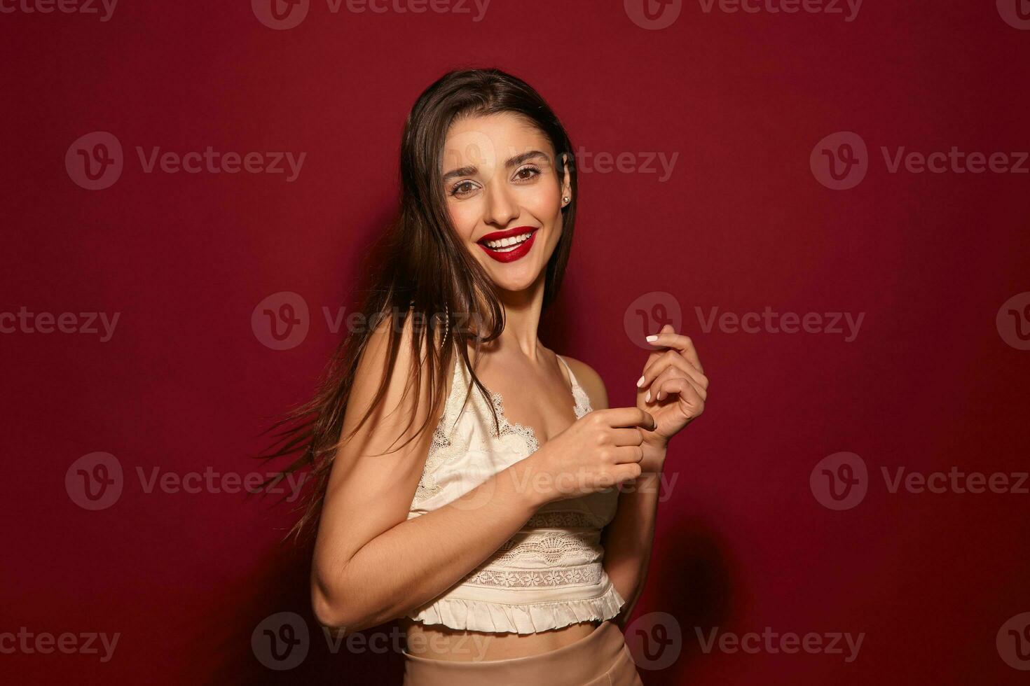 Cheerful young brown-eyed long haired brunette woman with festive makeup looking happily at camera with broad smile and keeping her hands raised while posing over burgundy background photo