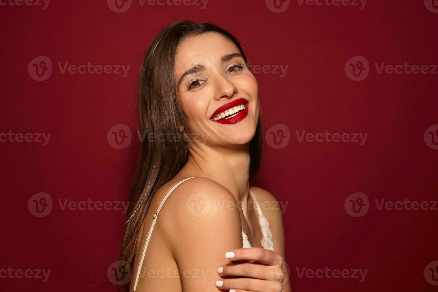 Portrait of young beautiful brown-eyed long haired brunette lady with red lips showing her white perfect teeth while smiling happily at camera, isolated over burgundy background photo