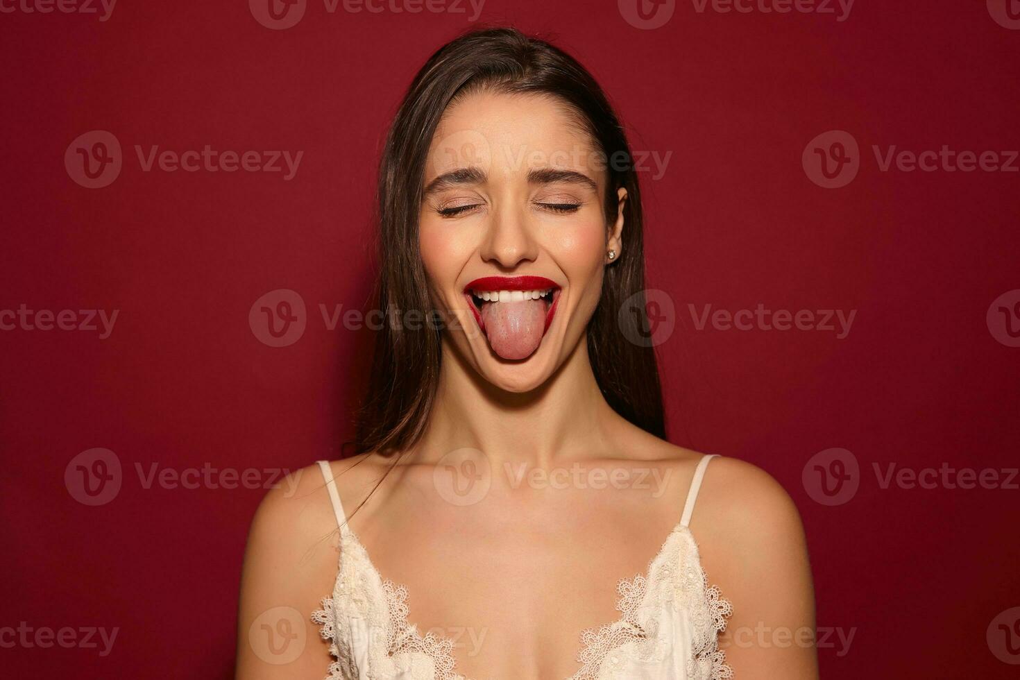 Good looking young attractive dark haired female with evening makeup sticking cheerfully out her tongue and keeping eyes closed, isolated over burgundy background photo