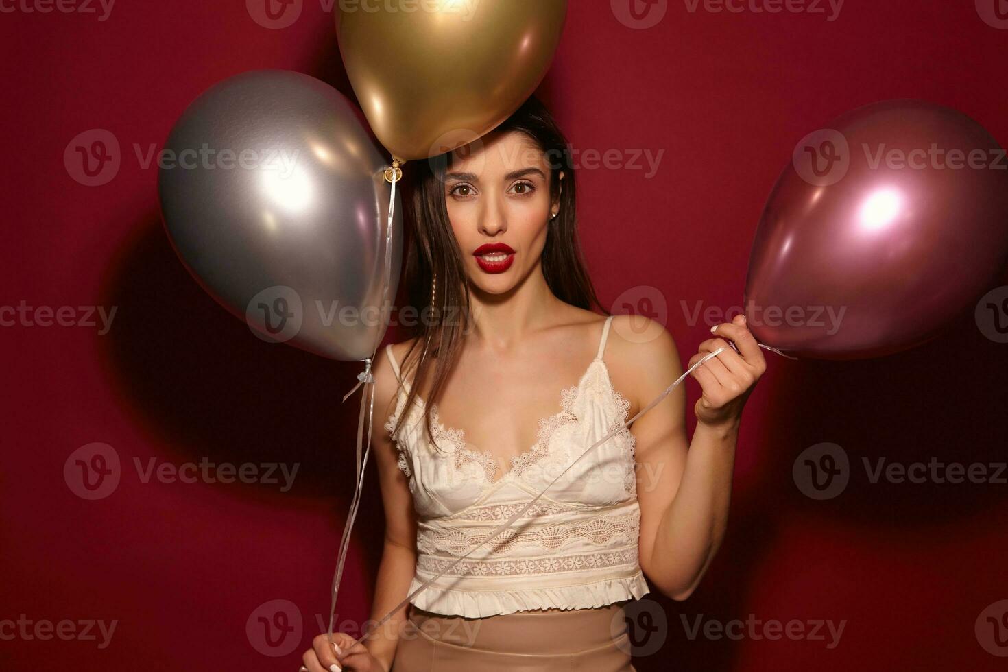 Studio shot of young brown-eyed long haired brunette woman with evening makeup holding air balloons and looking seriously at camera, isolated over burgundy background photo