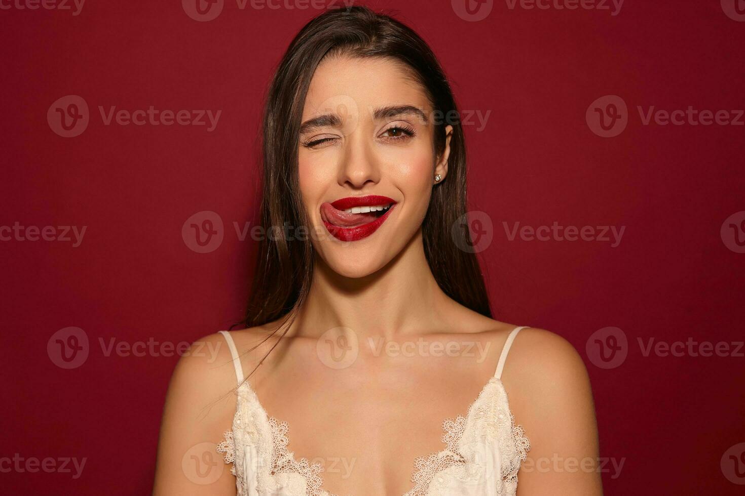 coqueta joven encantador largo peludo morena dama vestido en elegante ropa mientras posando terminado borgoña fondo, mirando alegremente a cámara con uno ojo cerrado y demostración su lengua foto