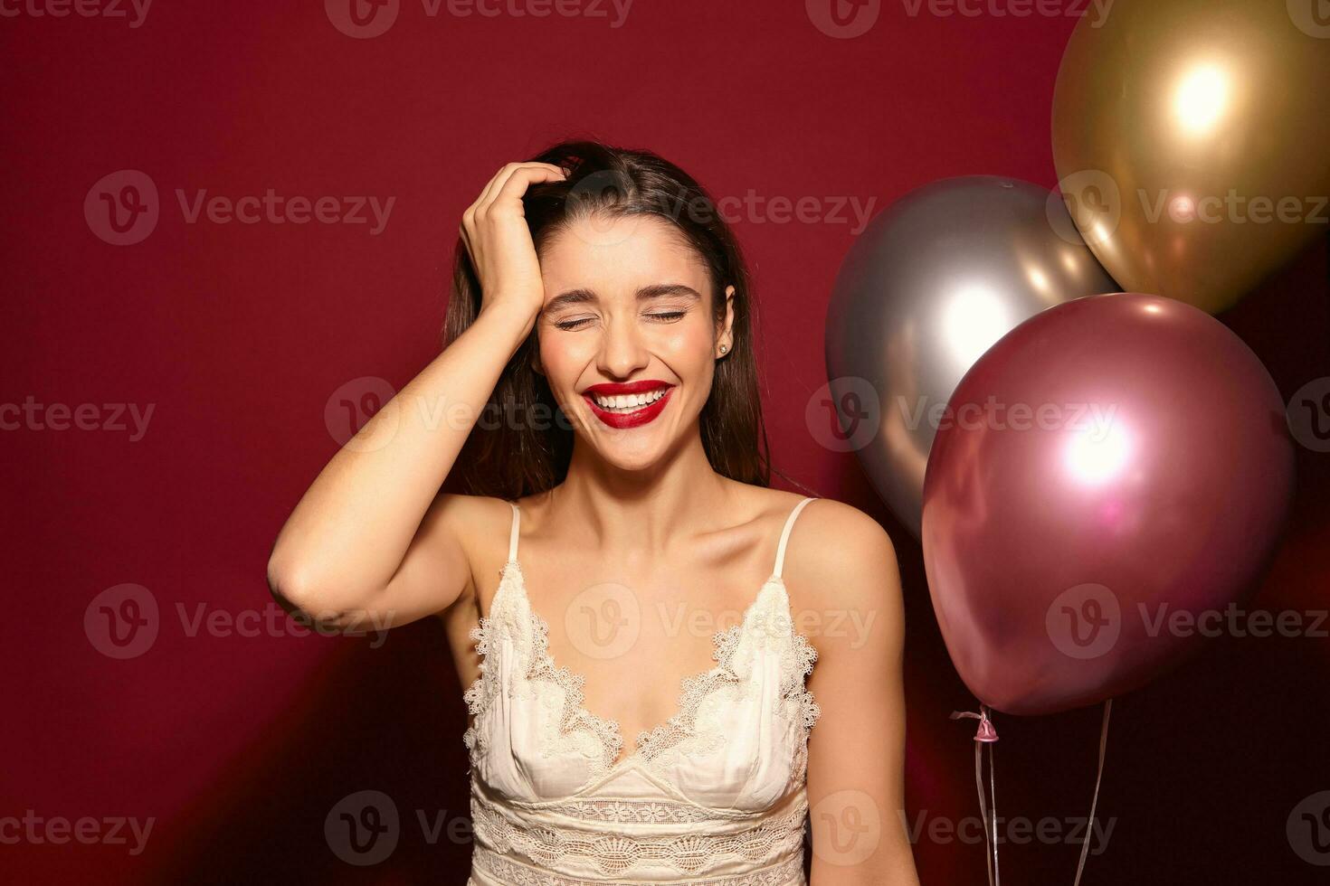 Happy young pretty brunette lady with festive makeup keeping her eyes closed while smiling cheerfully, being surprised with her friends and feeling amazed while posing over burgundy background photo