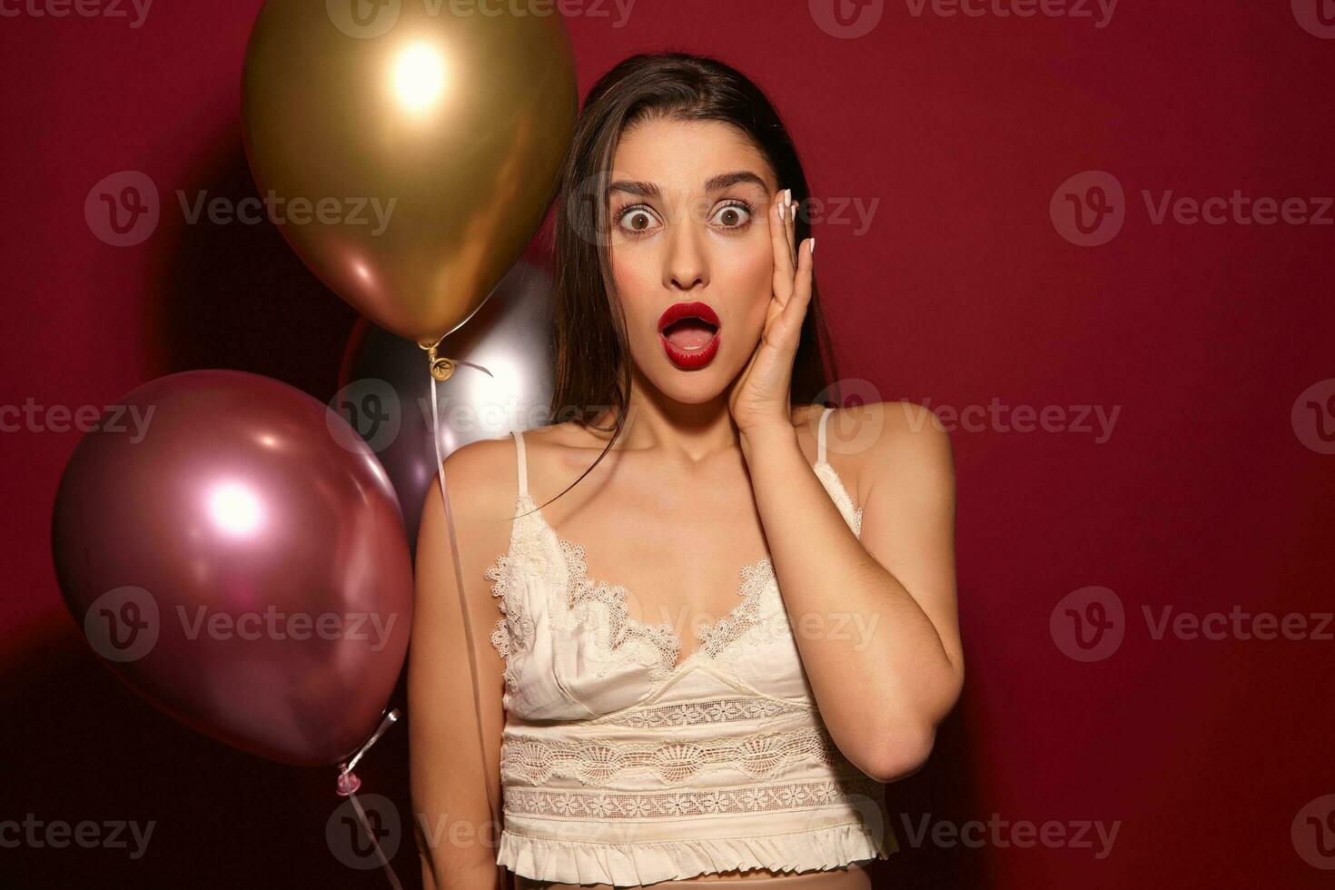 Shocked young beautiful dark haired elegant woman rounding her brown eyes while looking amazedly at camera and raising hand to her face, posing over burgundy background photo