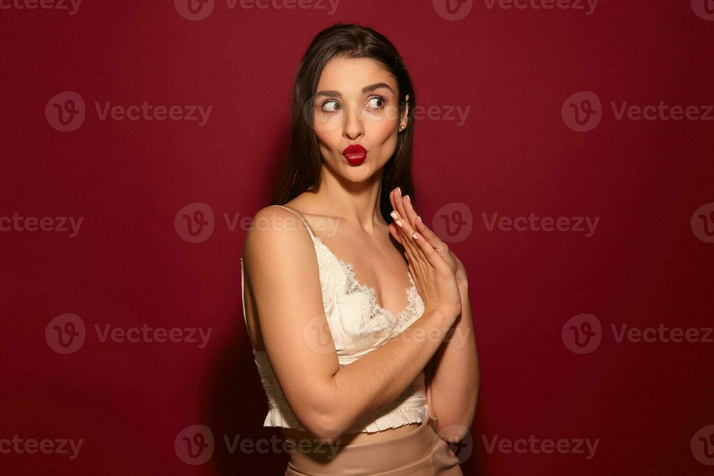 Studio shot of young lovely dark haired female with festive makeup pursing her lips and looking excitedly aside, keeping raised hands folded while standing over burgundy background photo