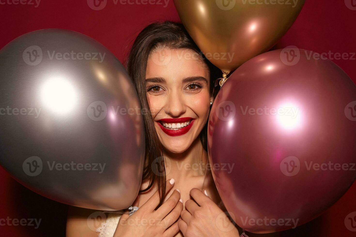 Portrait of joyful young brown-eyed brunette lady with red lips posing over colored air balloons while having nice party with her friends, being in high spirit and smiling broadly photo