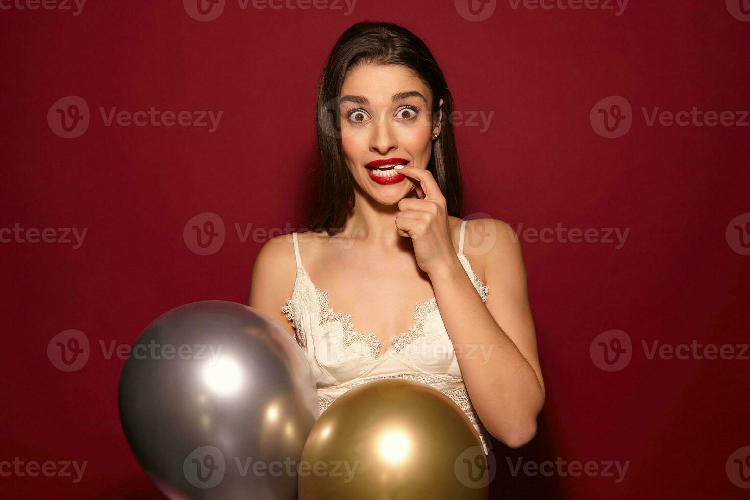 Open-eyed young attractive long haired brunette lady dressed in festive wear holding raised forefinger on her lip and looking amazedly at camera, isolated over burgundy background photo