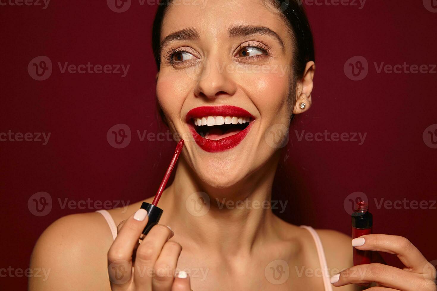 Joyful young attractive brunette woman with ponytail hairstyle looking cheerfully aside and smiling widely, preparing for going out and being in high spirit, isolated over burgundy background photo