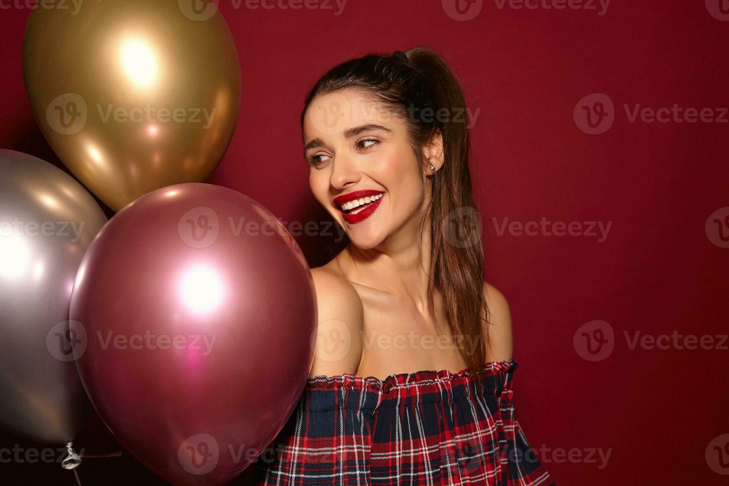 Cheerful young attractive dark haired lady with ponytail hairstyle being in nice mood while celebrating her birthday with friends, isolated over burgundy background photo