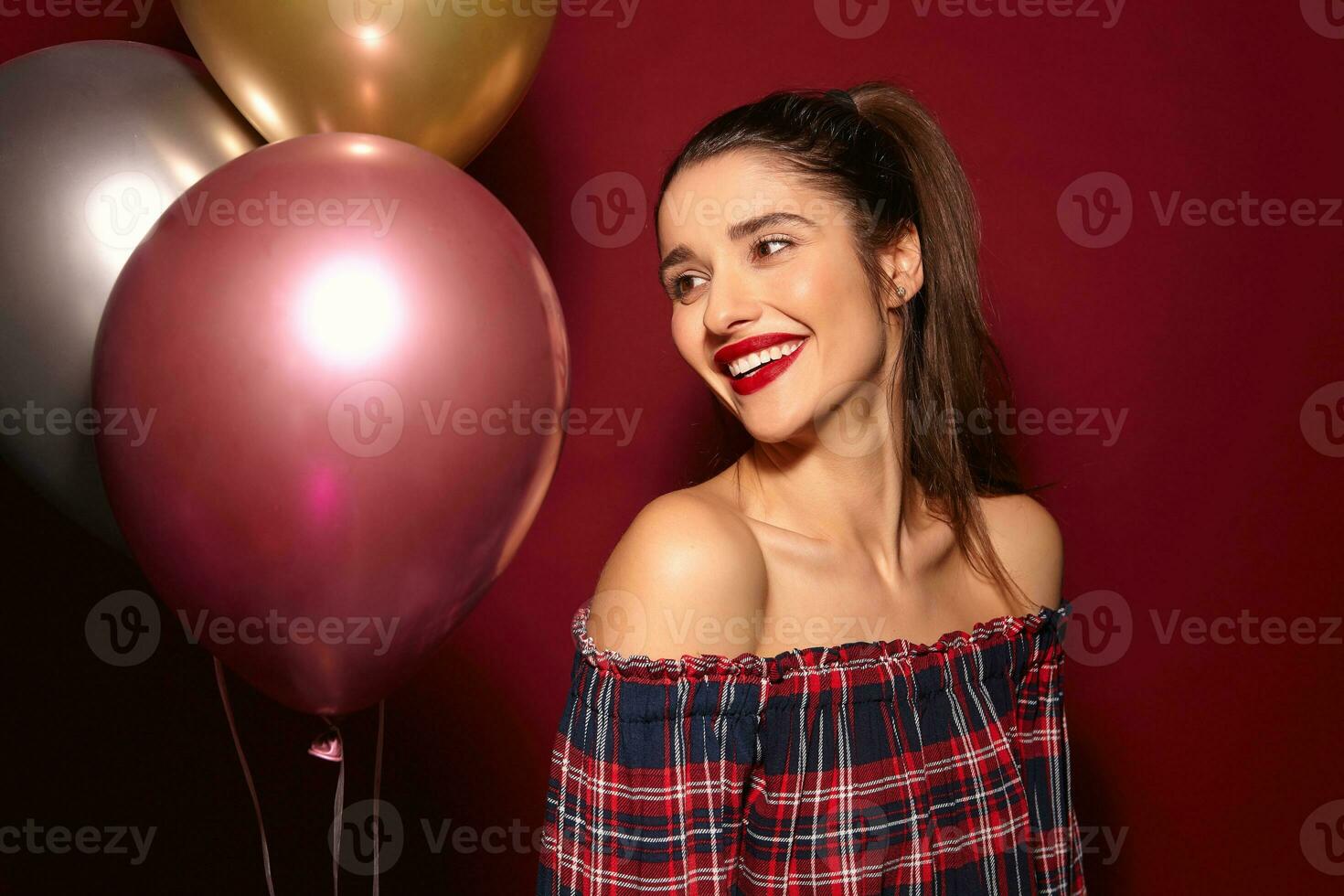 retrato de joven encantador de ojos marrones morena mujer con noche maquillaje sonriente agradablemente mientras posando terminado borgoña antecedentes con multicolor helio globos foto