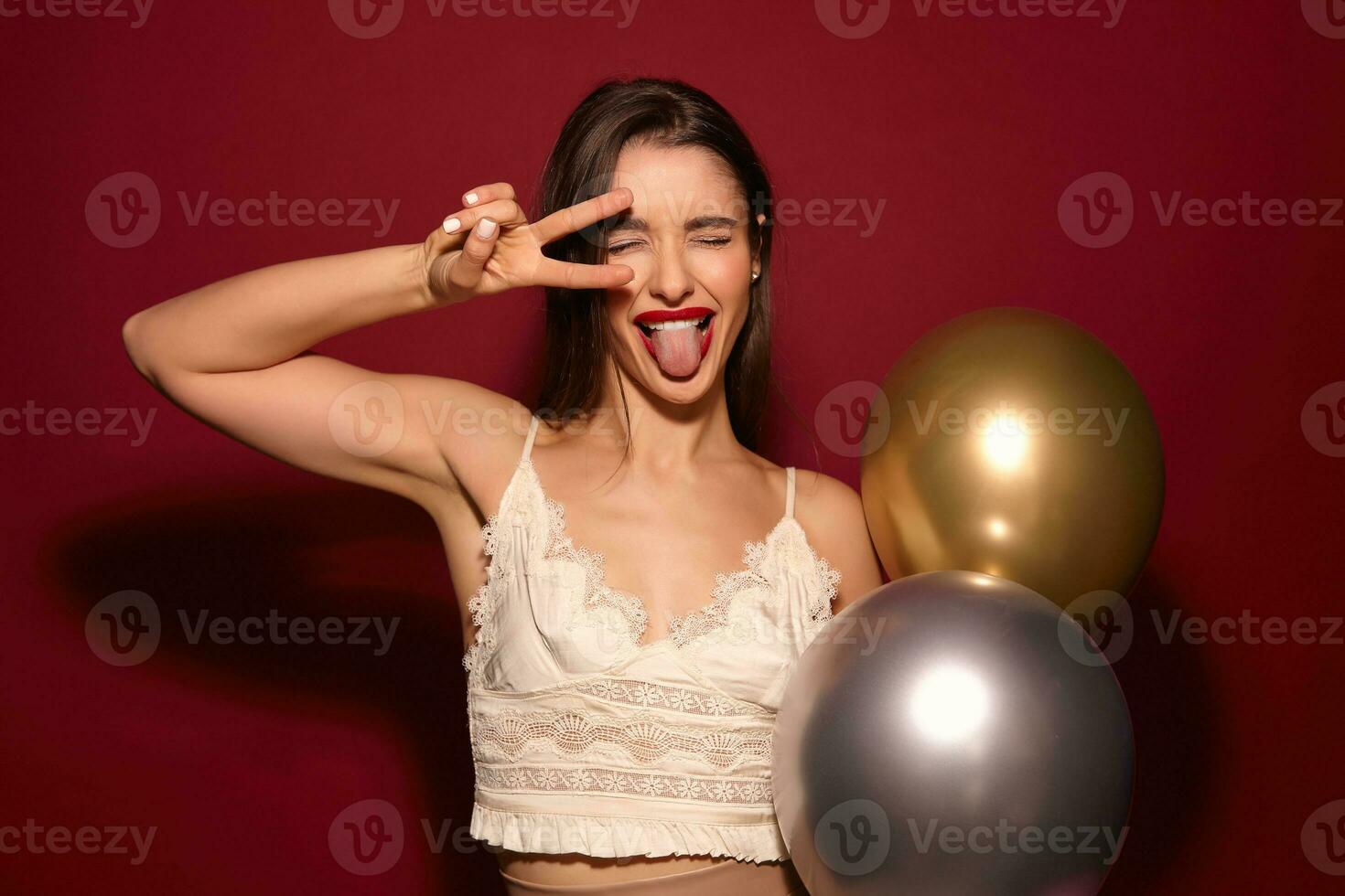 Portrait of young cheerful brunette female with evening makeup fooling while posing over burgundy background with air baloons, raising victory sign to her face and showing tongue photo