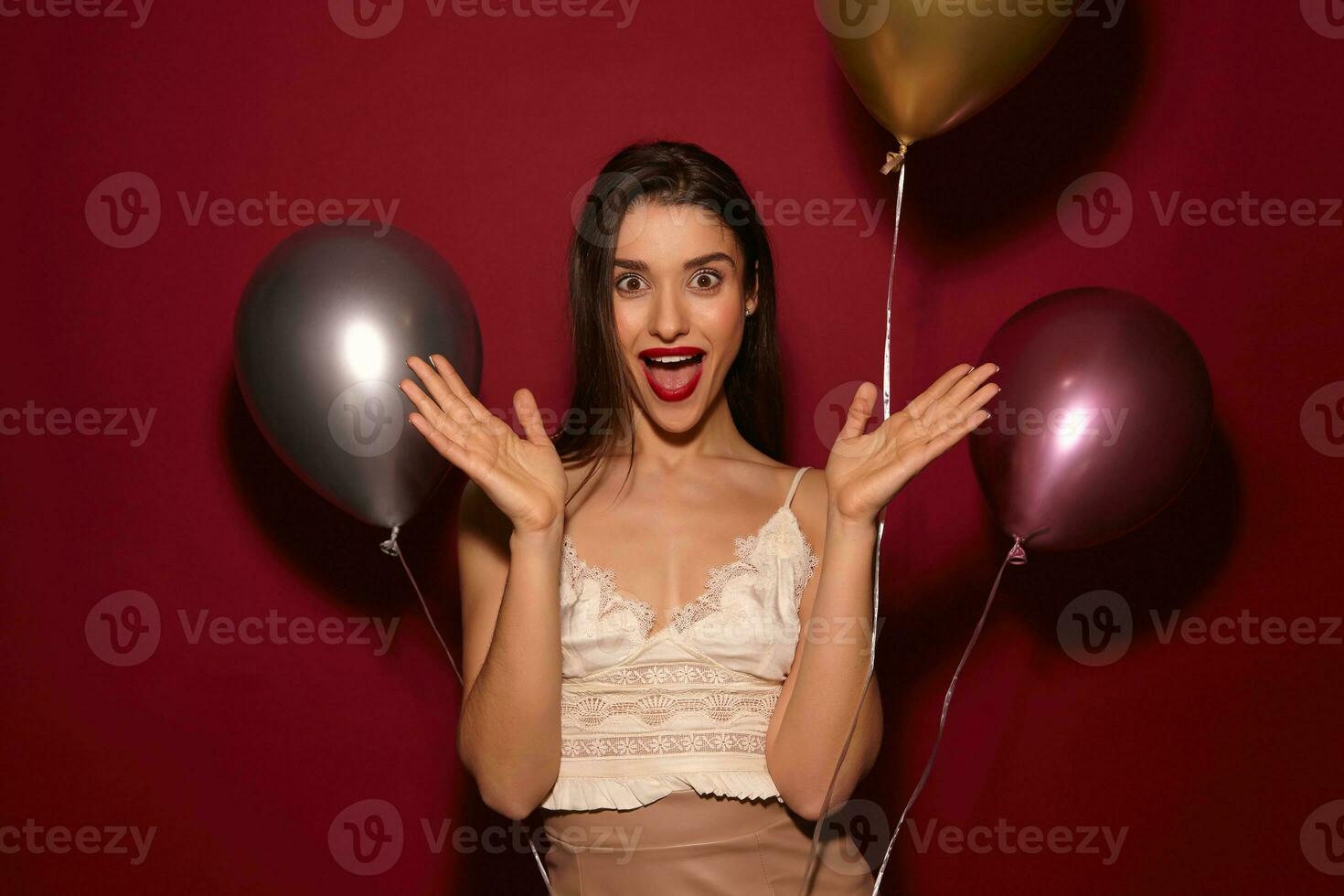 Astonished young beautiful brunette female with evening makeup raising emotionally her palms and looking at camera with wide eyes and mouth opened, isolated over burgundy background photo