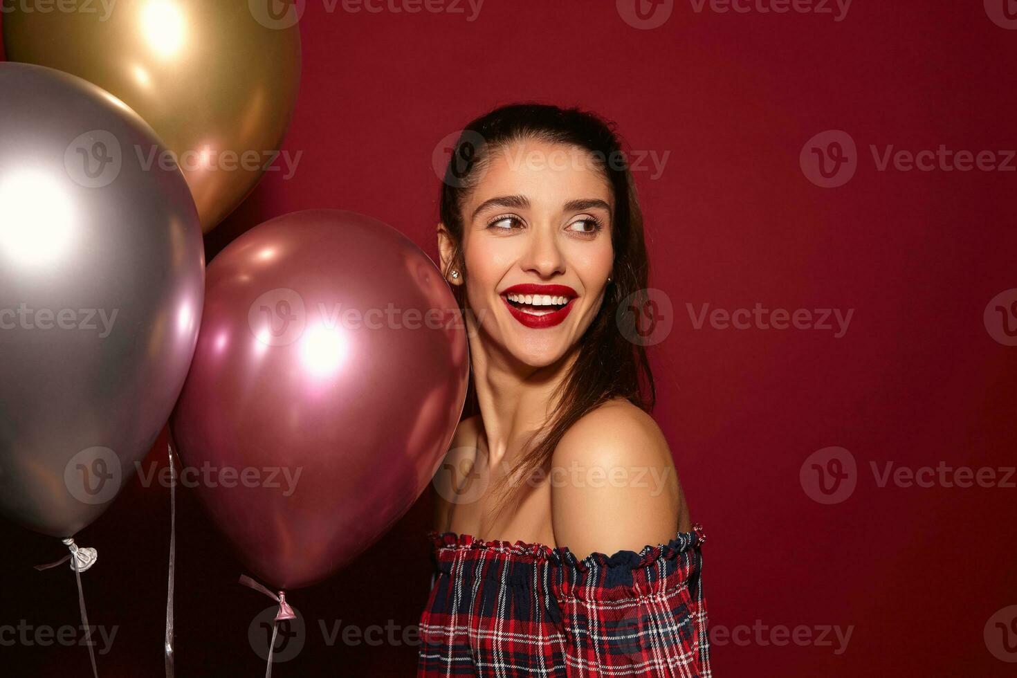 Happy young pretty dark haired woman with festive makeup looking cheerfully aside with charming smile while posing over burgundy background with air balloons photo