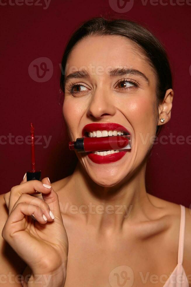 Portrait of attractive young brunette elegant woman fooling while making makeup and holding lip gloss with her mouth, posing over burgundy background in strap top photo