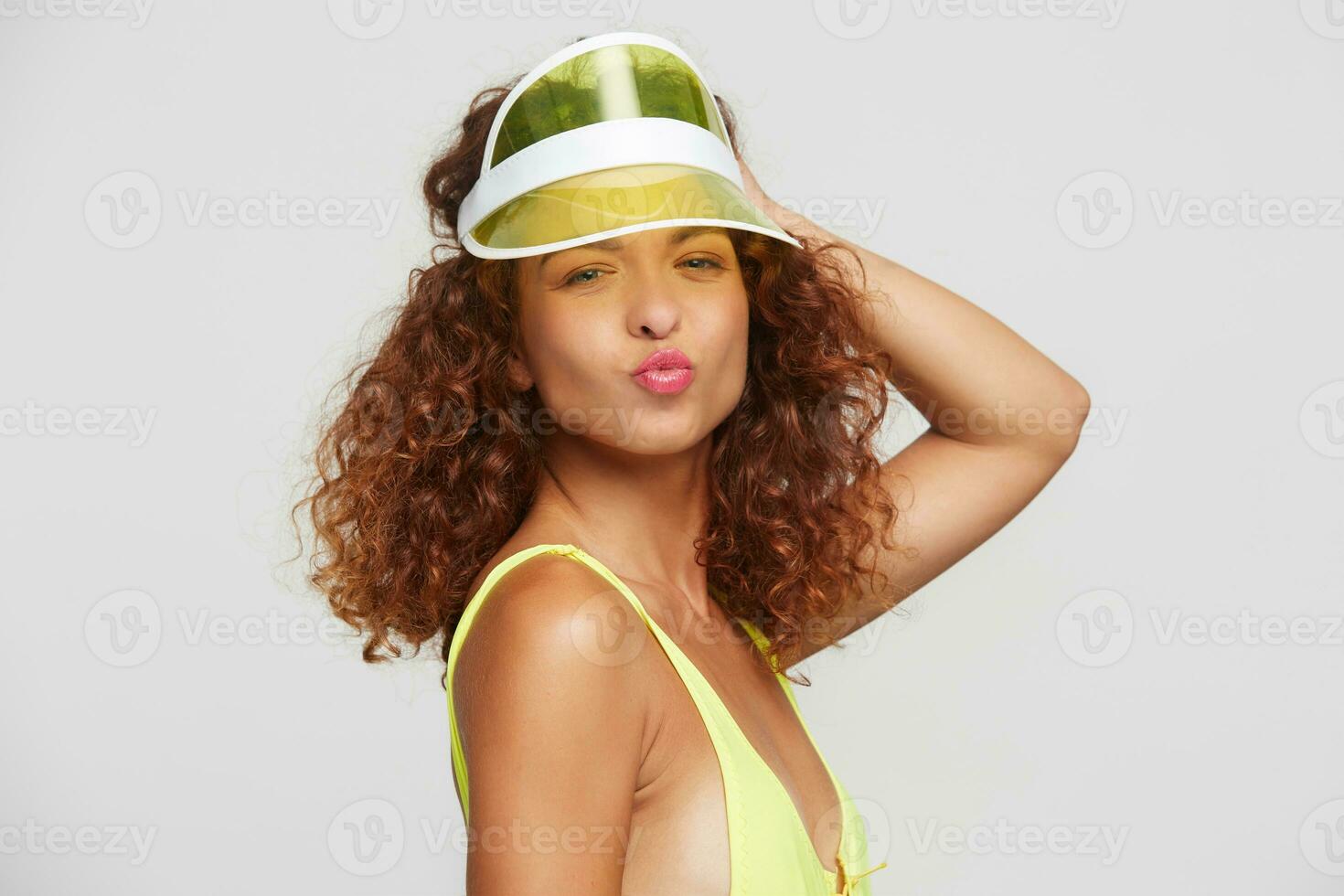 Positive young pretty redhead curly lady folding lips in air kiss and raising hand to her neon cap while standing over white background, going to beach party with friends photo