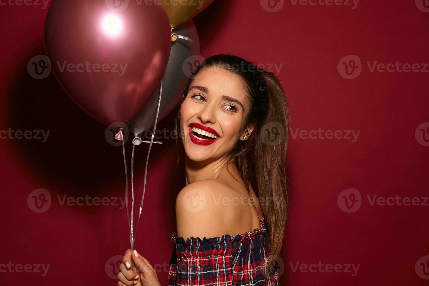 Joyful young beautiful dark haired female with evening makeup keeping air balloons in raised hand and smiling widely, wearing elegant top while posing over burgundy background photo