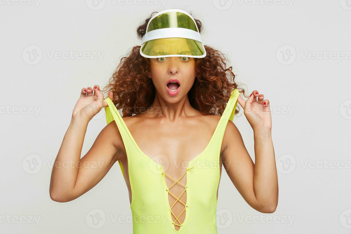 Bewildered young green-eyed redhead curly female with casual hairstyle pulling her swimming suit with raised hands and looking confusedly at camera, isolated over white background photo
