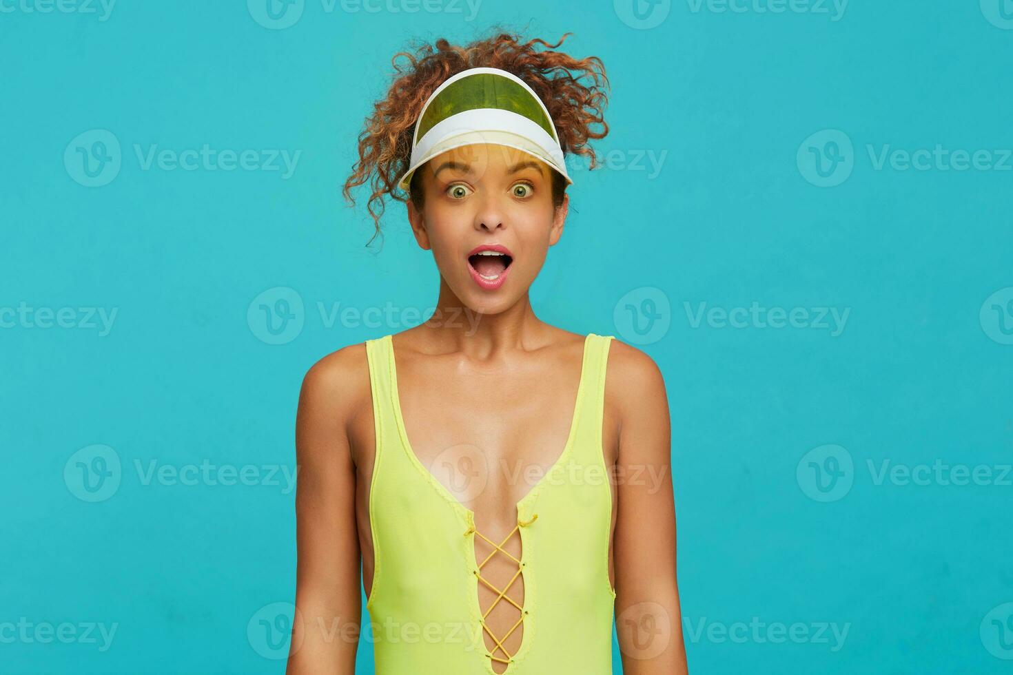 Bemused young pretty redhead curly lady looking amazedly at camera with wide eyes and mouth opened, dressed in swimming wear while standing over blue background photo