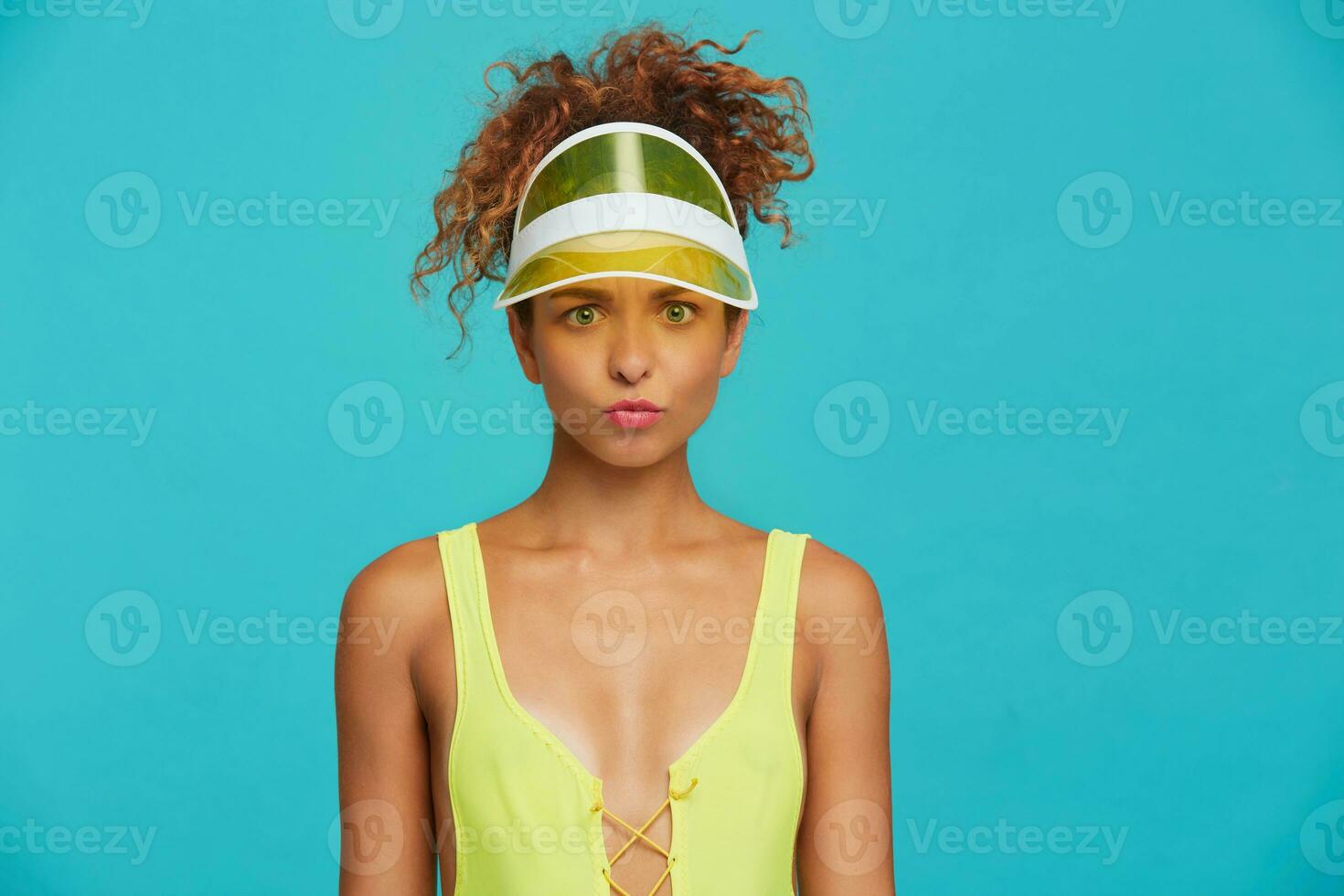 Portrait of confused young pretty redhead curly lady with ponytail hairstyle looking at camera with pout while standing over blue background with hands down photo