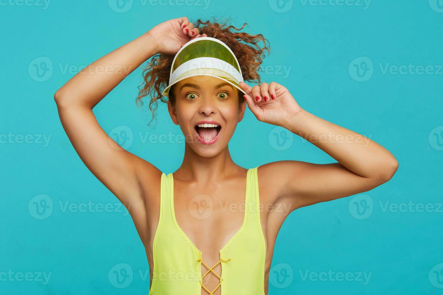 Agitated young lovely red haired woman with ponytail hairstyle looking excitedly at camera with wide eyes and mouth opened while posing over blue background with raised hands photo