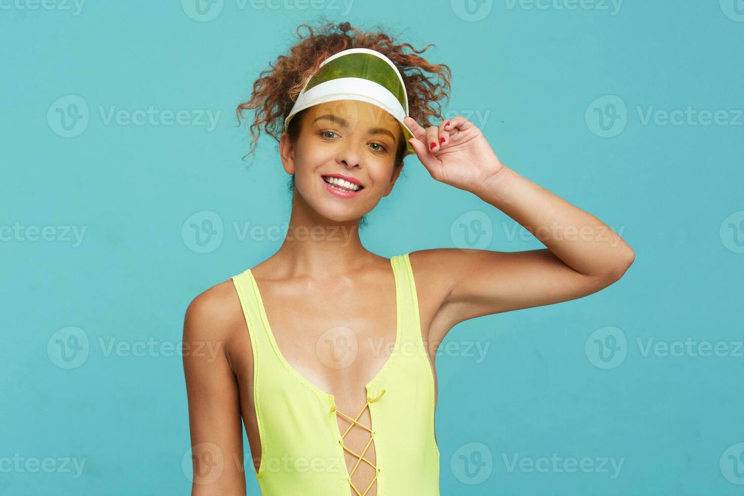 Glad young attractive slim redhead woman with casual hairstyle holding her cap with raised hand and smiling cheerfully at camera while posing over blue background photo