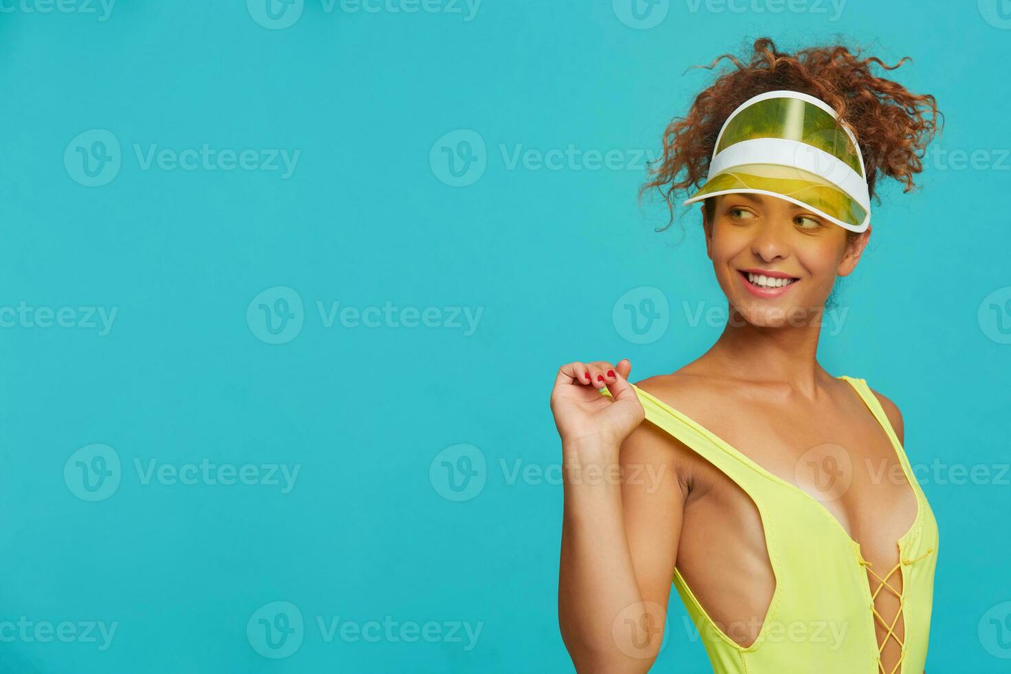 Pleased young attractive redhead curly female smiling widely while looking over her shoulder and keeping hands on swimsuit, isolated over blue background photo