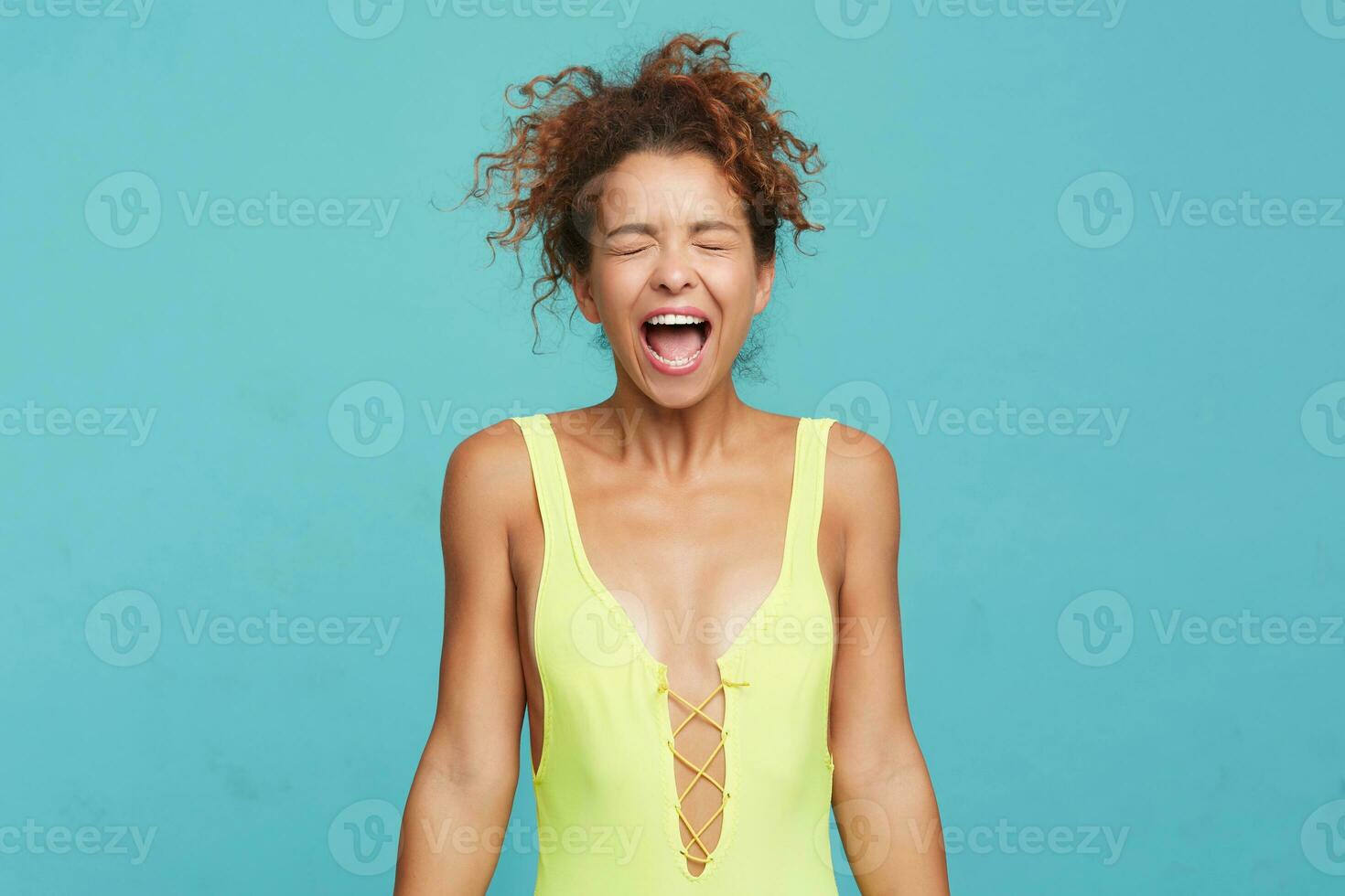 Excited young attractive curly woman with ponytail hairstyle keeping her hands closed while screaming with wide mouth opened, isolated over blue background photo