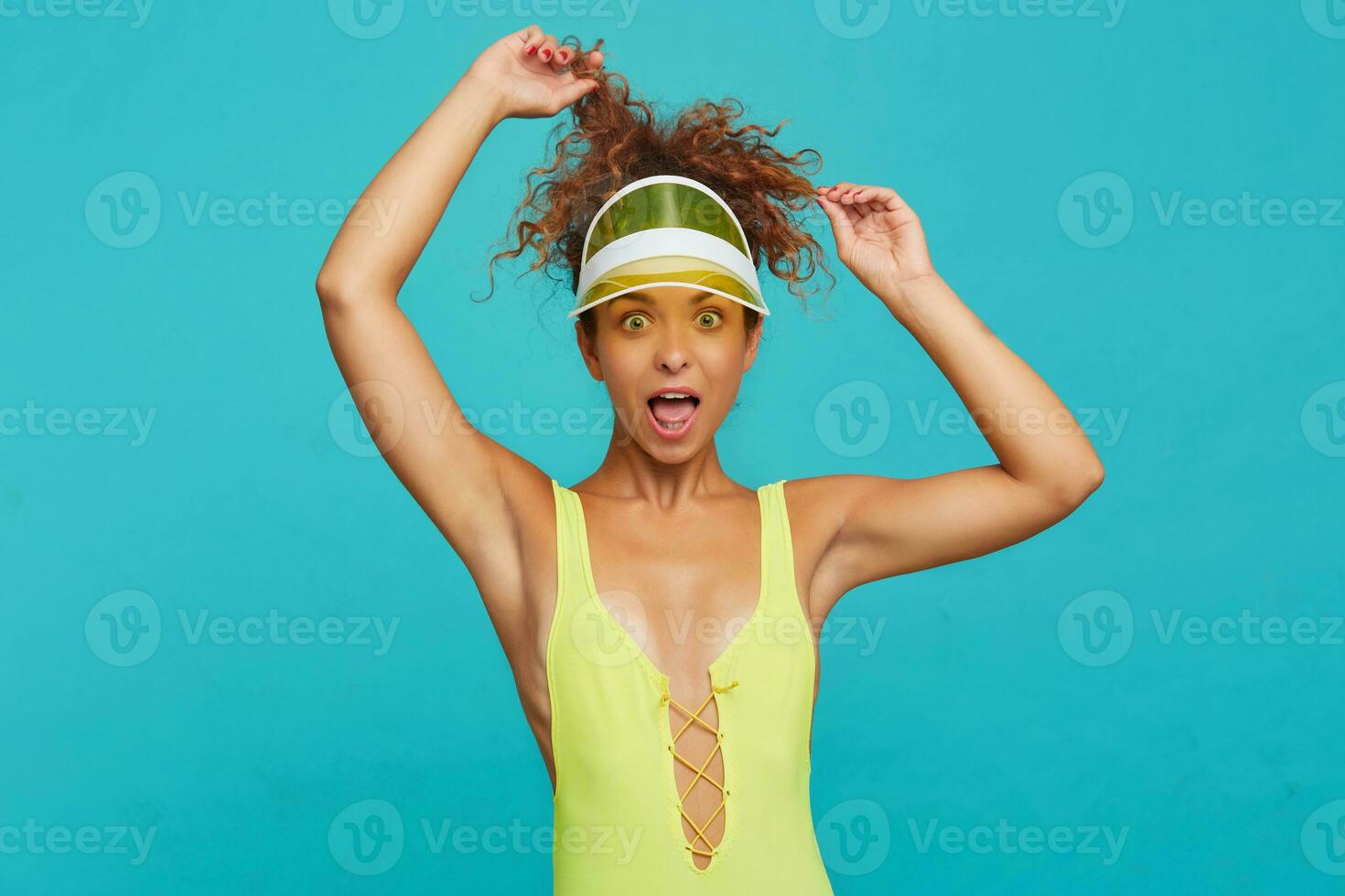 Excited young attractive curly woman with natural makeup keeping raised hands above head while looking amazedly at camera with wide eyes opened, isolated over blue background photo