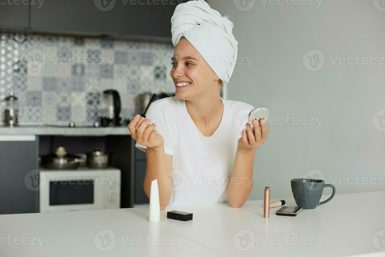Portrait of a beautiful girl making make-up in the morning, drinking tea, looking and smiling to someone a side, dressed casually and head wrapped in a towel, on a kitchen background photo