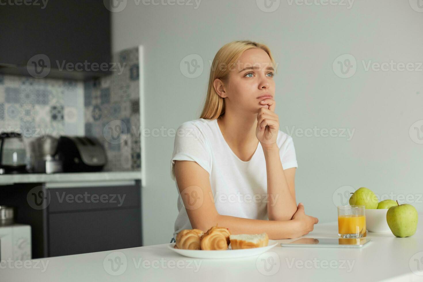 A young blonde girl sits at the kitchen table, sad, looks thoughtfully to the window side, on the table -a glass of juice, tablet, apples, buns, dressed in a domestic white T-shirt, kitchen background photo