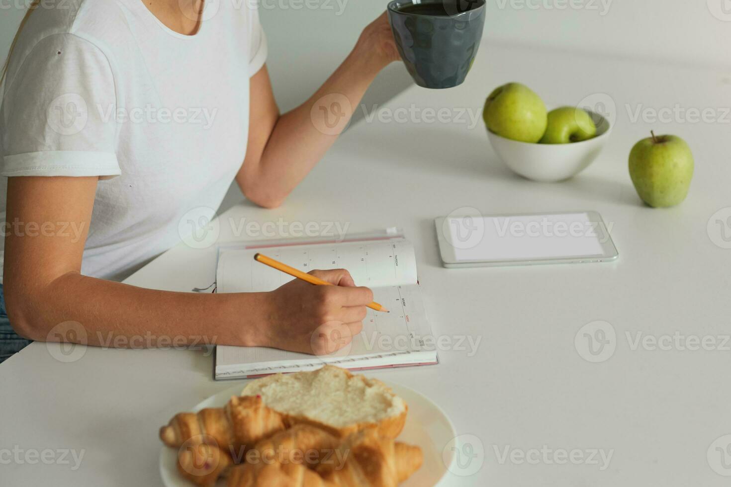 Close up woman hands, view from a side, planning her day, keeps a grey cup with some liquid, Croissants on a plate, apples, phone with white screen for your text are there on a white kitchen table photo