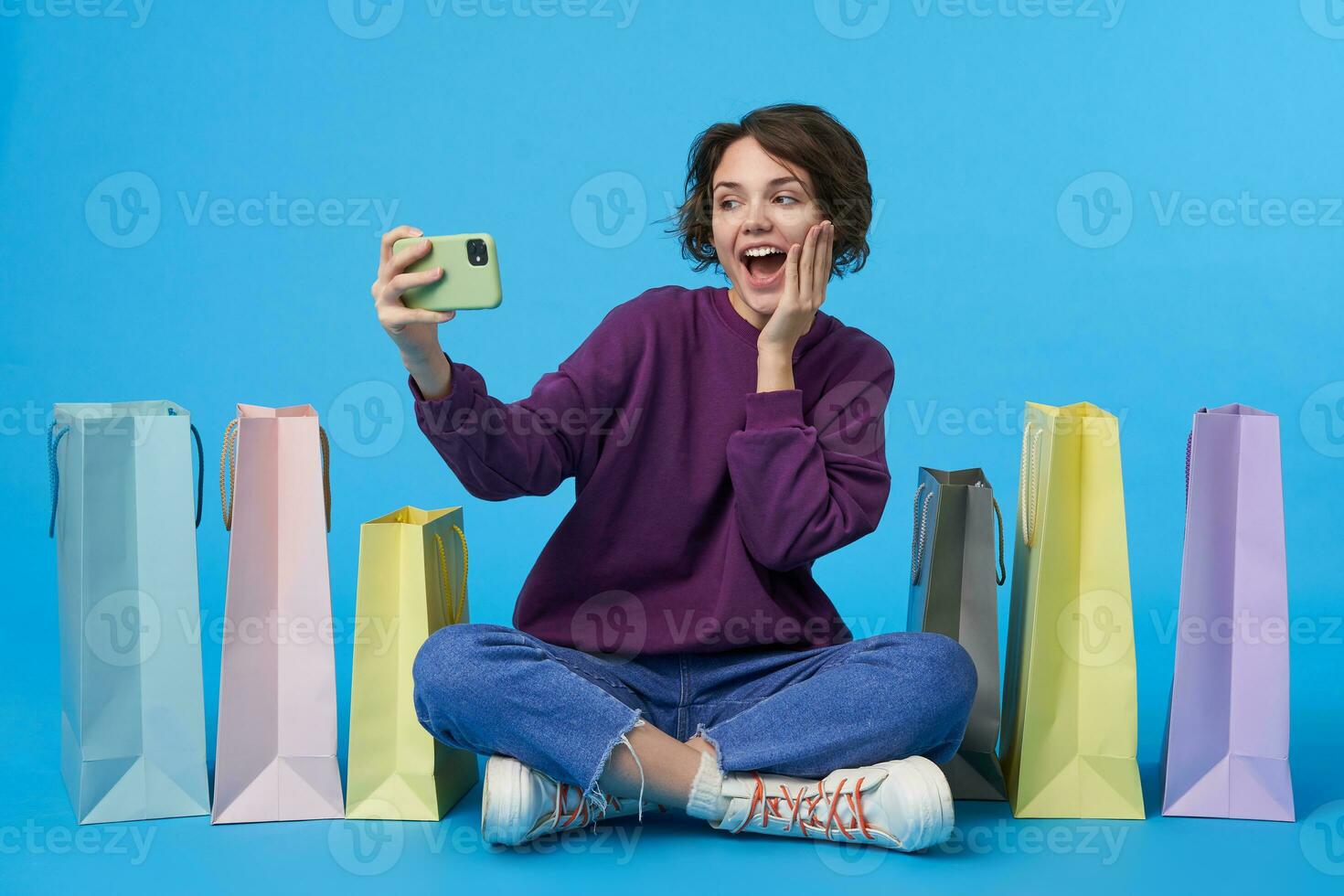 Cheerful young attractive short haired curly woman raising hand with mobile phone while making selfie and looking gladly at camera, sitting over blue background with crossed legs photo