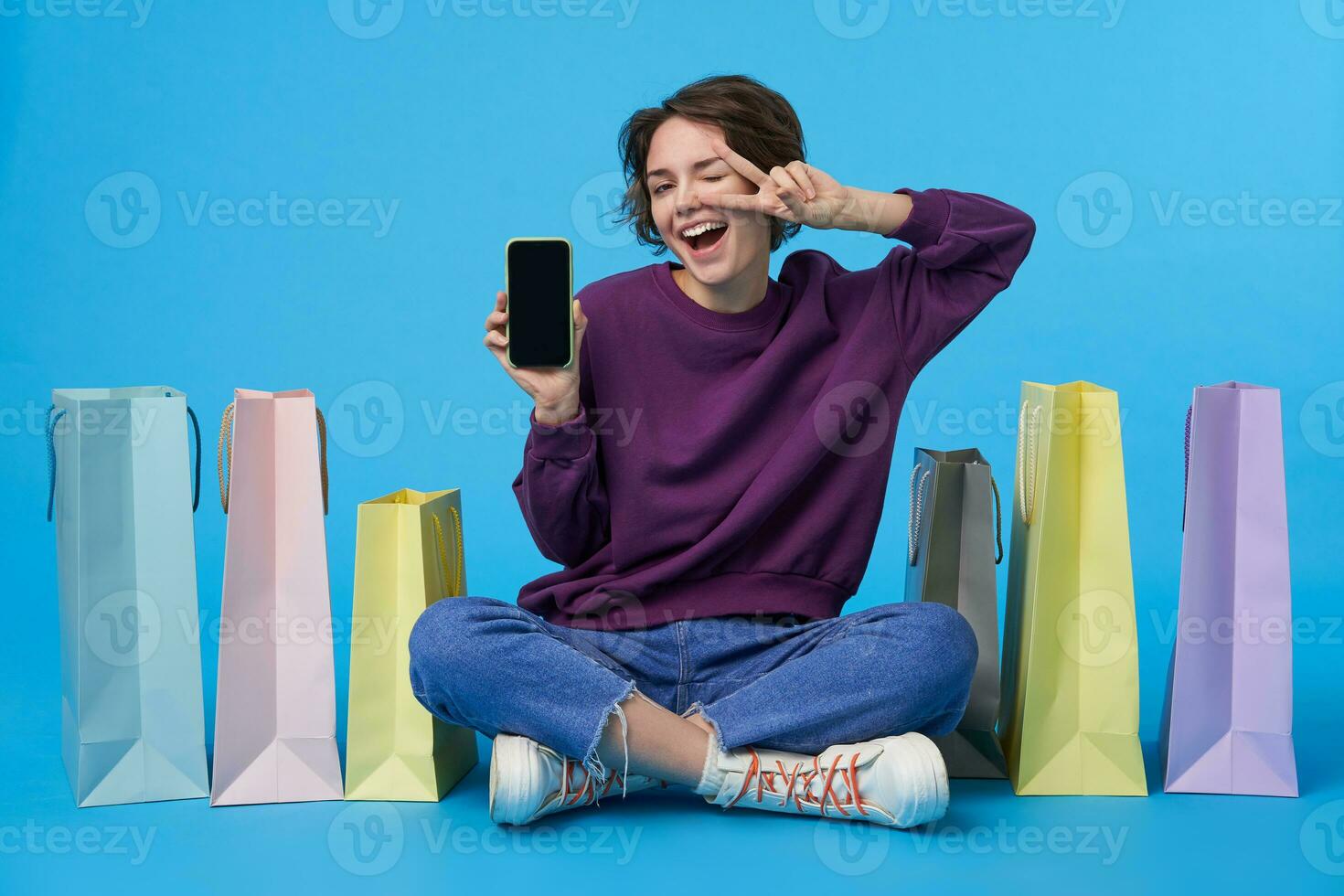 Cheerful young pretty dark haired curly female showing screen of her phone to camera and raising hand with victory gesture, sitting over blue background with paper bags photo