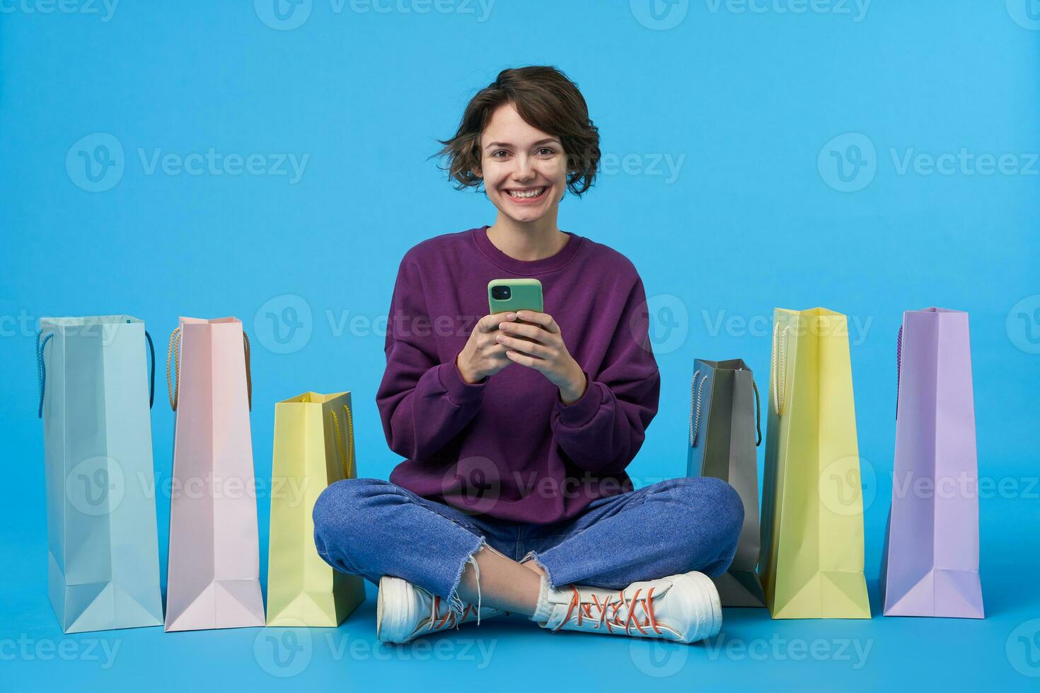 Cheerful young pretty curly brunette woman with casual hairstyle keeping smartphone in raised hands and looking happily at camera, sitting over blue background with crossed legs photo